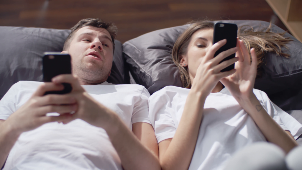 young-couple-lying-together-on-their-bed-using-their-mobile-phones-and-talking_rdx9gicm8__F0000